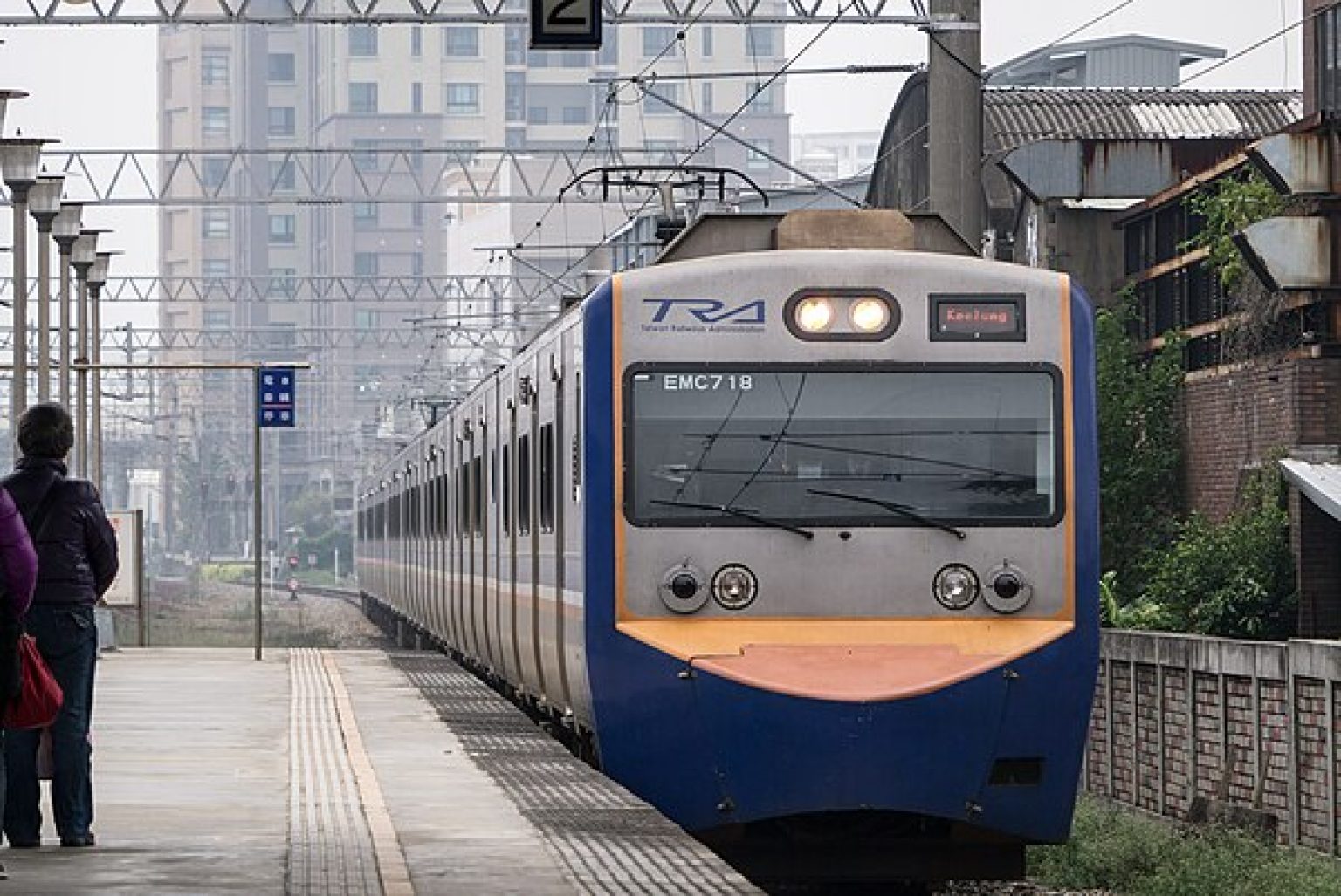 Taipei rail transportation structure and train with city in the background, suburban homes sit next to rail line, person stands waiting to get on train